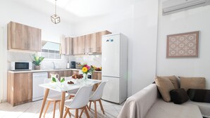 A modern kitchen with wooden cabinets, a white refrigerator, a dining table with chairs, and a living area with a sofa. - Family friendly holiday home  (Neo Kalamaki)