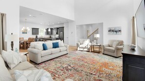 A spacious living room with a large sofa and armchairs, a colorful rug, and a kitchen in the background.