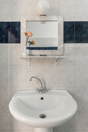 Un baño con lavamanos blanco, un espejo encima y un jarrón con flores en una repisa.