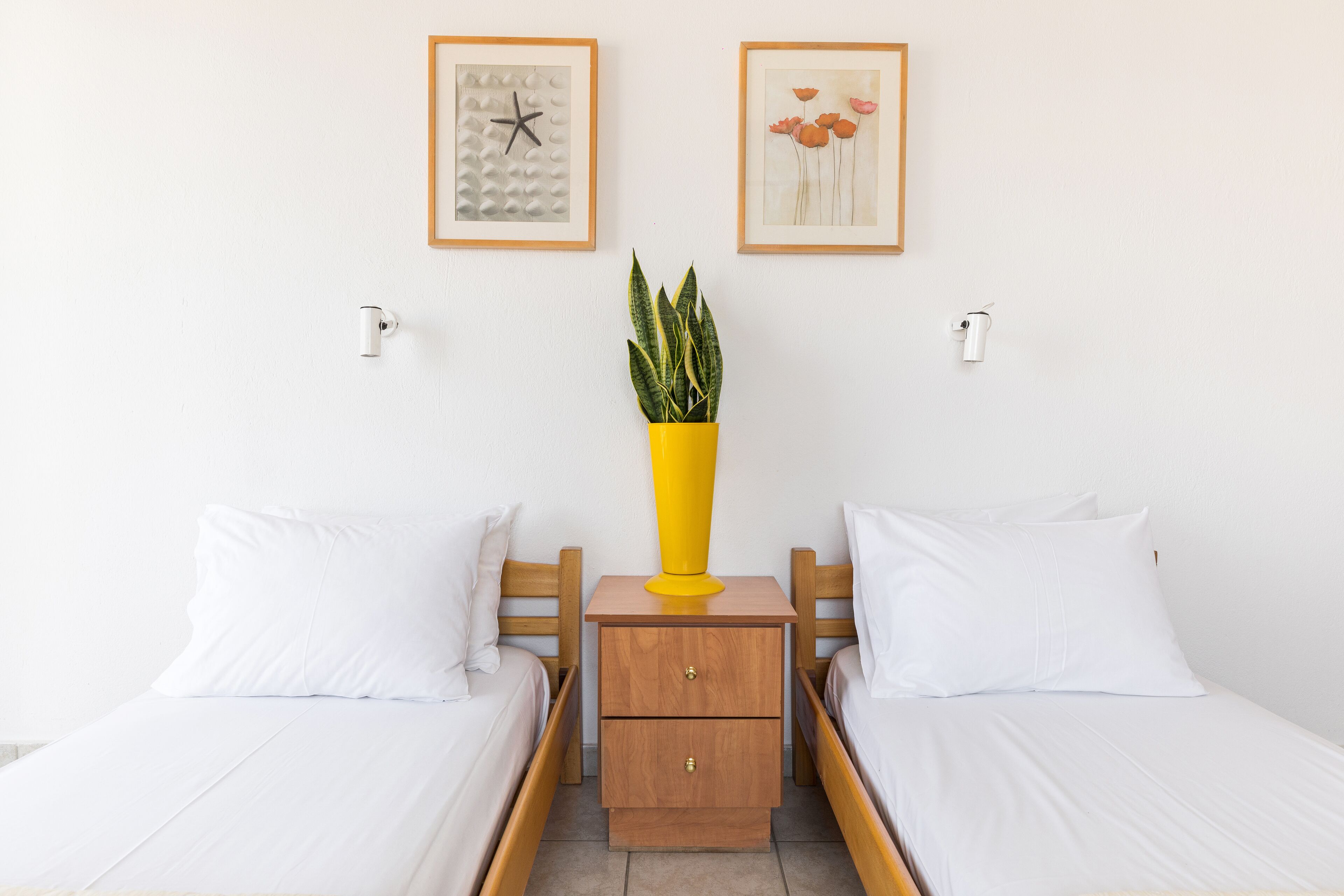 Una habitación de hotel con dos camas individuales, una mesita de noche de madera con un jarrón amarillo y dos cuadros en la pared.