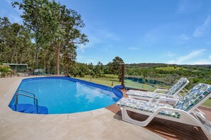 Ein Schwimmbecken mit Holzterrasse und Liegestühlen, umgeben von Bäumen und Blick auf eine grüne Landschaft.