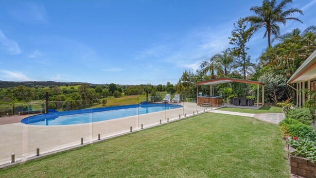 Una piscina con un porche techado, rodeada de un césped cuidado y plantas tropicales.