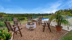 Un patio con sillas de madera y un hogar para fuego, con vista a una piscina y abundante follaje.