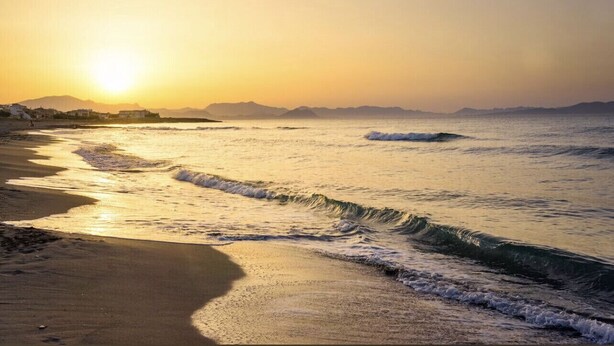 Ein Strand mit Wellen im Abendrot, ein klarer Himmel und ferne Berge.