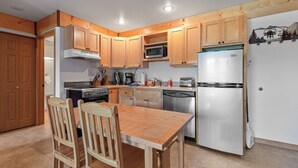 A compact kitchen with wooden cabinets, a stainless steel refrigerator, and a dining area with wooden chairs. - Ski in Ski out at Schweitzer Mountain Resort | Great Views | Cozy Condo (Sandpoint)