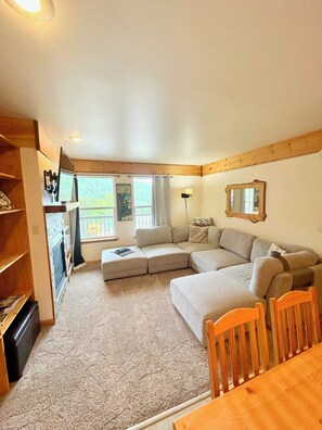 A cozy living room with a sectional sofa, wooden beams, a TV mounted on the wall, and a view of the outdoors.