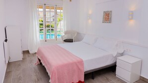 A bedroom with a bed, a nightstand, a chair, a window with curtains, and a view of a pool. - Isla Saint Tropez Apartment Javea Arenal, with Pool, Wifi, AC and recently Refurbished (Javea / Xàbia)