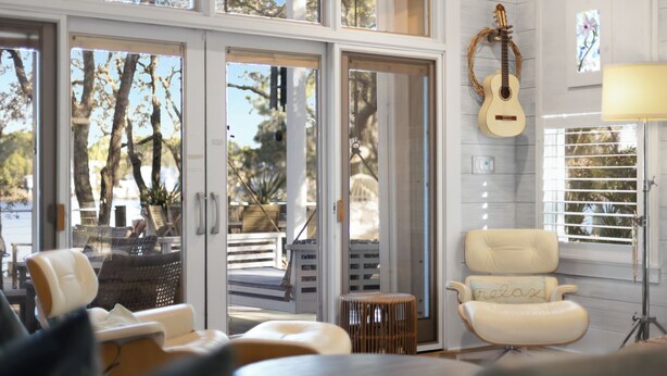 Un espacio habitable amplio con un gran ventanal corredera que da a una terraza exterior, un sillón beige cómodo, una pequeña mesa redonda y una guitarra en la pared.