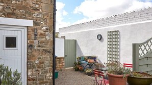 Un edificio de piedra con una puerta blanca y una ventana pequeña. Hay plantas en macetas y un carretilla con herramientas de jardinería.