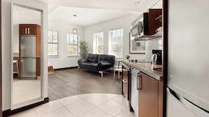 A modern kitchen with a refrigerator, microwave, and stove. There is a black leather sofa in a living area with a view of the outdoors.