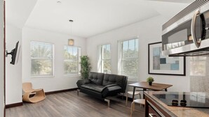 A modern kitchen with a built-in microwave, a dining area with a table and chairs, a black leather sofa, and large windows with blinds.