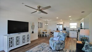 A modern living room with a flat-screen TV, a ceiling fan, a patterned armchair, and a dining area with a wooden table and chairs. - SHORE BEATS WORK IS 500 STEPS TO BEACH! MODERN, IMMACULATE TOWNHOME! (Mexico Beach)