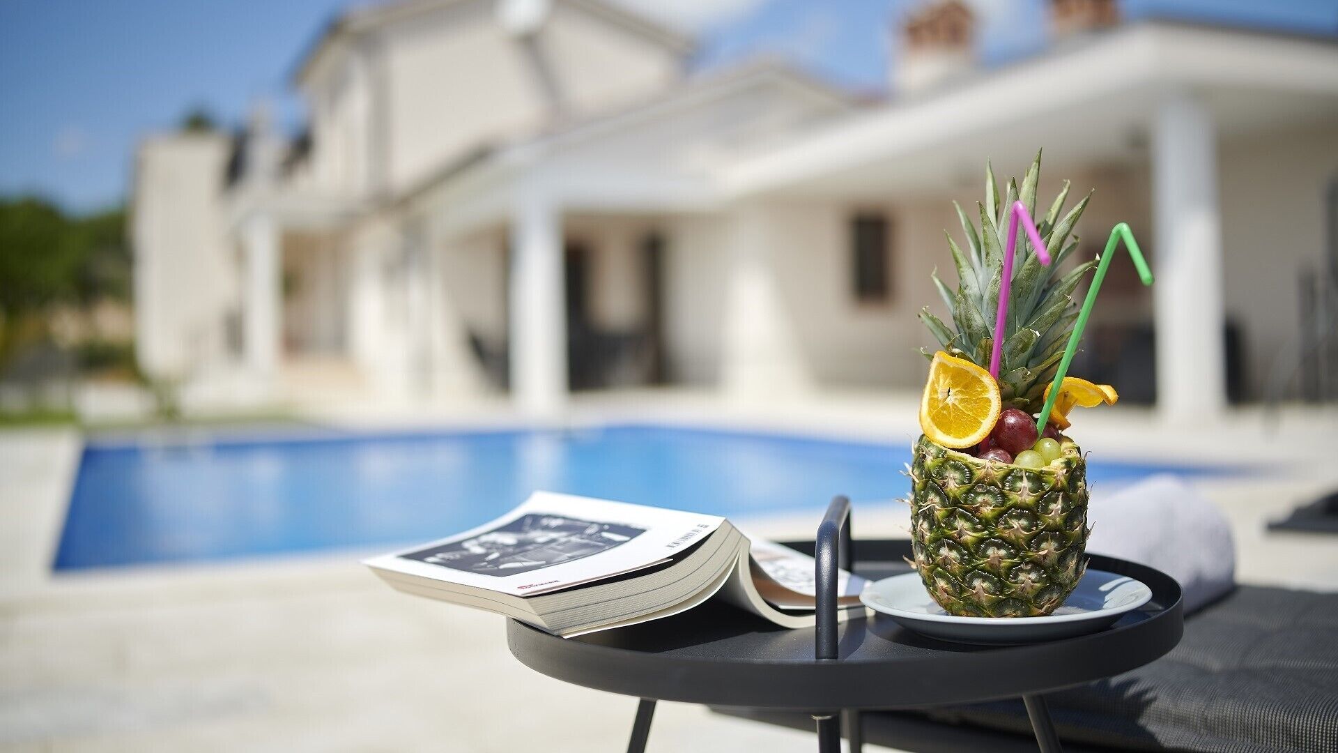 Een ananascocktail op een tafel met een tijdschrift, in de buurt van een zwembad en een modern huis op de achtergrond.