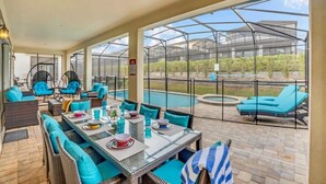 Un patio cubierto con una mesa lista para comer, sillas y un área de descanso junto a la piscina.