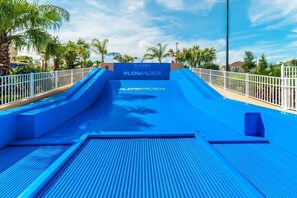 Um escorregador azul com a marca 'FlowRider', rodeado por uma cerca branca e palmeiras.