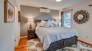 A bedroom with a bed, bedside lamps, a wall-mounted air conditioner, a decorative mirror, and a window with curtains.