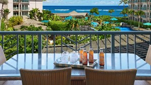 A balcony with a table set for two, overlooking a pool area with umbrellas and lounge chairs, and a view of the ocean.