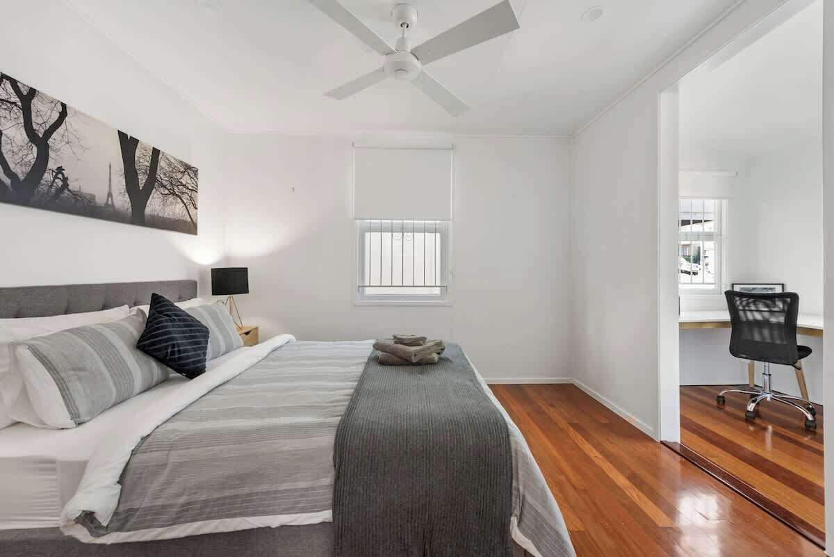 A bedroom with a bed, a ceiling fan, a window with blinds, a nightstand, a chair, and a painting on the wall.