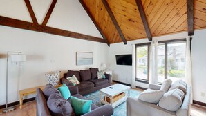 Sala de estar con techo de madera, un sofá gris, una mesa de centro blanca y vistas al exterior a través de amplios ventanales.