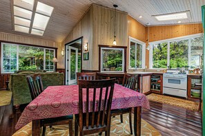 A cozy dining area with a wooden table and chairs, a kitchen with a stove, and large windows offering views of greenery.