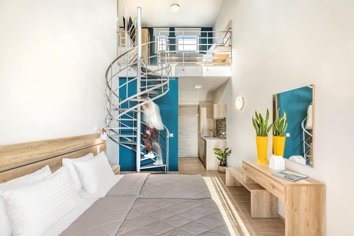A modern bedroom with a spiral staircase, a bed, a desk, and a view of the kitchen.