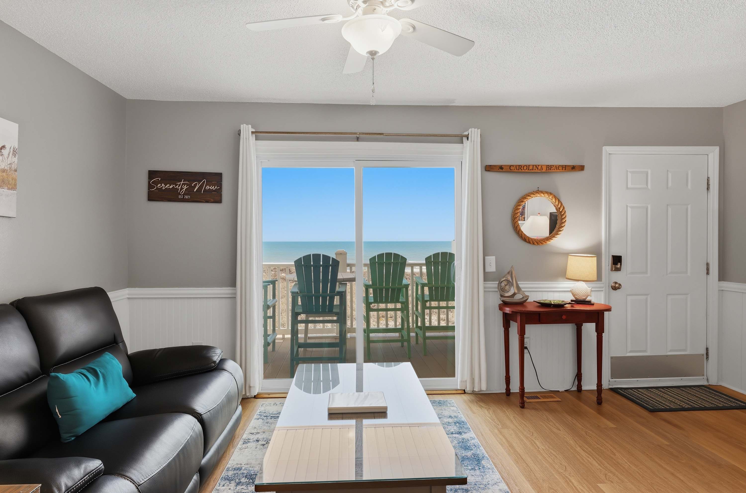 A living room with a leather sofa, a coffee table, a view of the ocean through a sliding glass door, a ceiling fan, and a small table with a lamp.