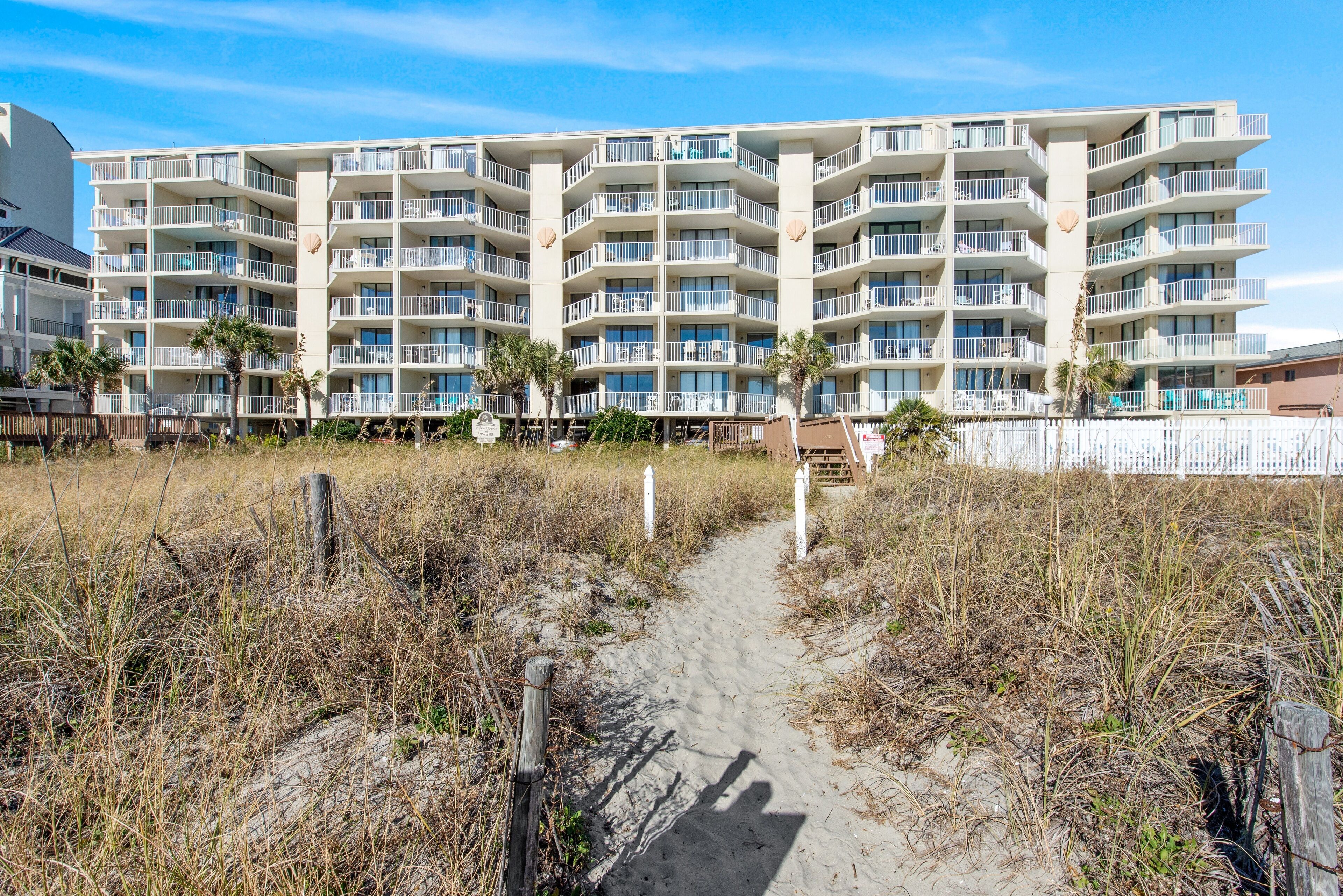 A beachfront hotel with multiple balconies, a sandy path leading to the entrance, and a white fence.