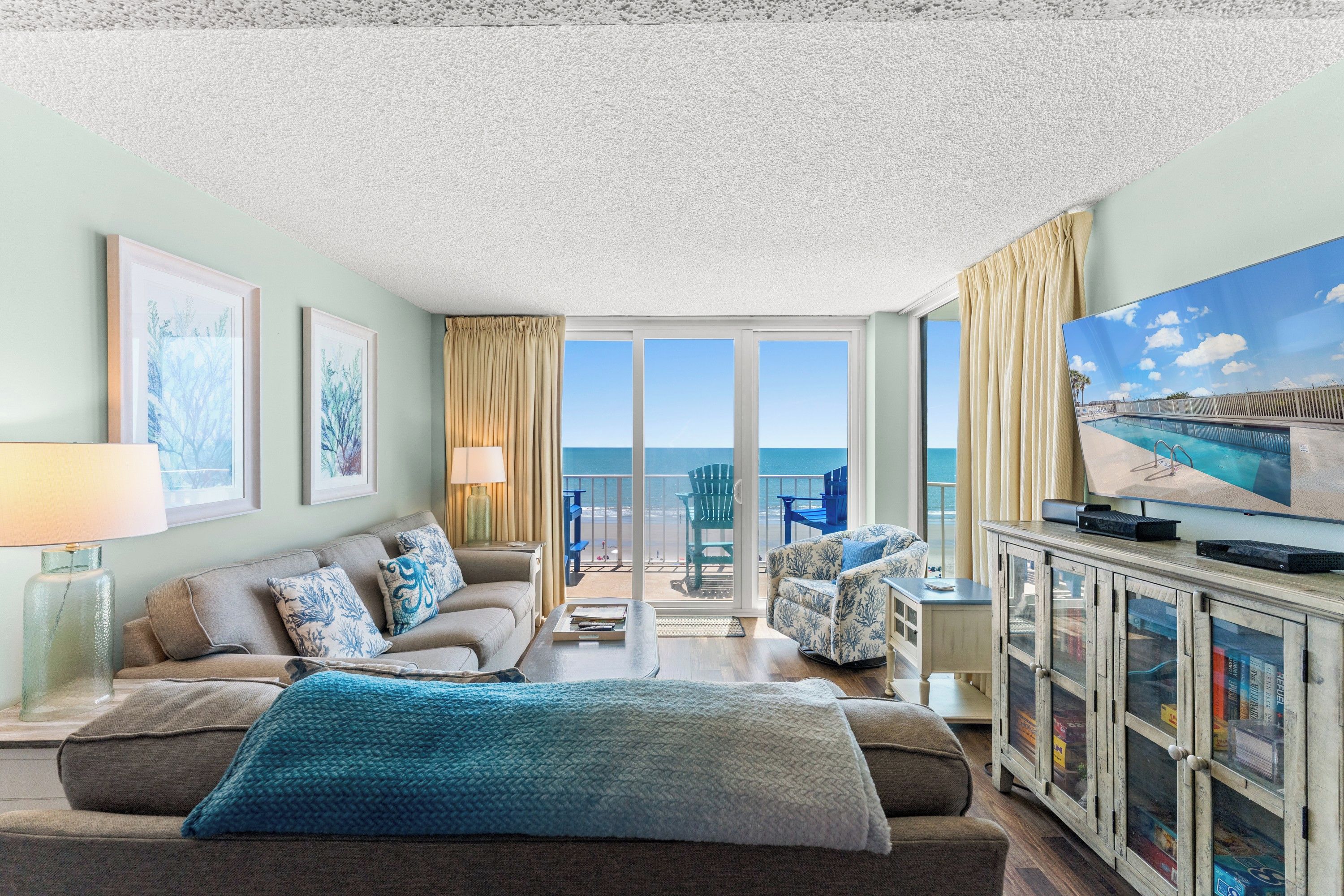 A spacious living room with a large television, a sofa, a coffee table, and a view of the ocean through the glass doors.