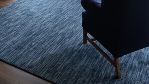 Un interior de madera con un ventilador de techo, una alfombra azul, un sillón morado, un sofá beige y un cuadro con un paisaje enmarcado.