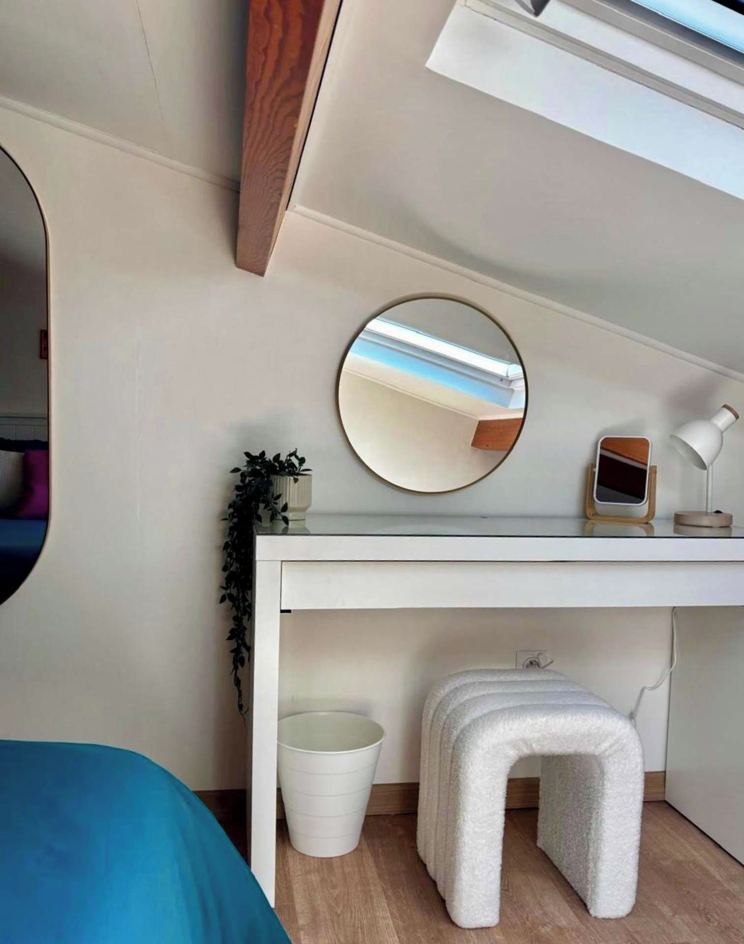 Ein modernes Zimmer mit weißem Schreibtisch, rundem Spiegel, kleinem Hocker, Topfpflanze und weißem Mülleimer.
