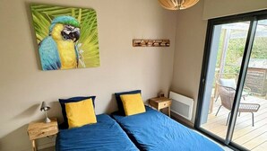 A bedroom with a large bed, two bedside tables, a lamp, a painting of a parrot, and a view of an outdoor deck. - Modernes Ferienhaus in Erster Reihe Gegenüber der Dünen (Hourtin)