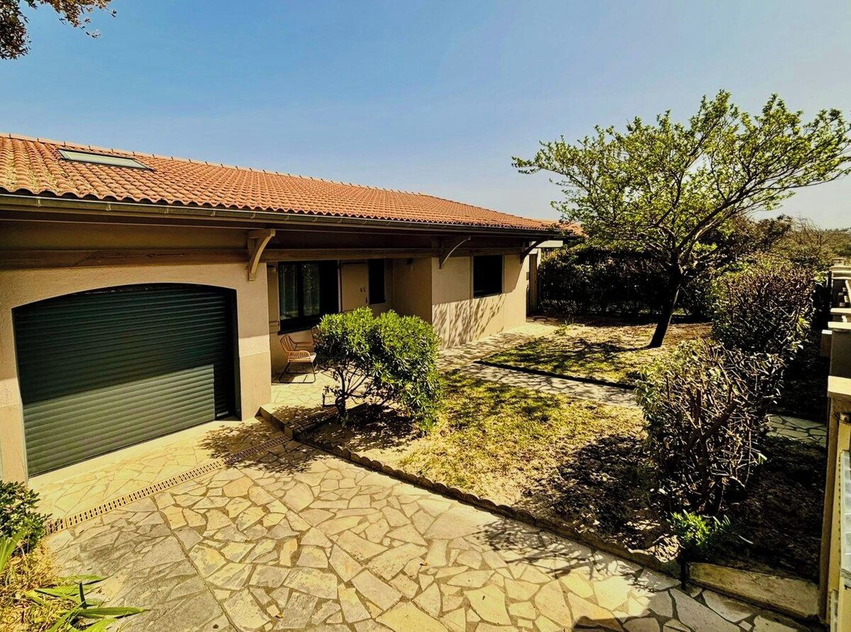 Une maison avec un toit en tuiles, un portail de garage, une allée pavée et un petit jardin agrémenté d’arbustes et d’un arbre.