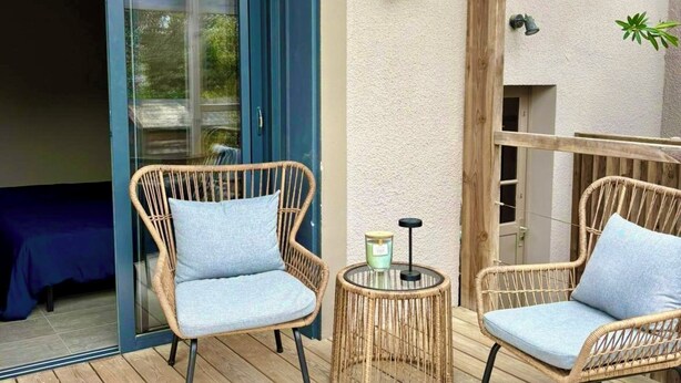 A wooden deck with wicker chairs and a small table, a glass door leading inside, and a textured wall. - Modernes Ferienhaus in Erster Reihe Gegenüber der Dünen (Hourtin)