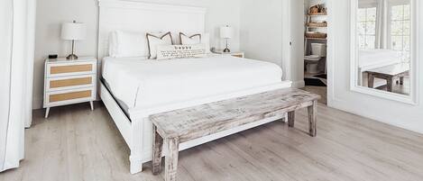 Un dormitorio con una cama grande, un banco de madera, un ventilador de techo, una mesita de noche con una lámpara y vistas al exterior por una ventana.