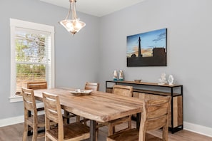 Uma sala de jantar com mesa e cadeiras de madeira, uma luminária pendente, um quadro na parede e uma janela com persianas.