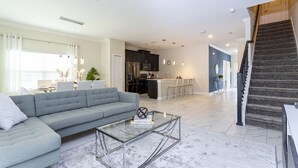 A modern living room with a grey sofa, a glass coffee table, and a kitchen in the background.