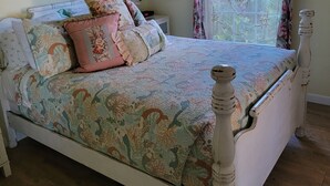 A bedroom with a bed, a ceiling fan, a window with floral curtains, and a wall-mounted air conditioner.