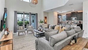 Ein geräumiges Wohnzimmer mit grauem Sofa, einem Couchtisch und einem Fernseher auf einem Holzboden. Im Hintergrund befindet sich eine Küche mit dunklen Schränken und einer Mikrowelle.