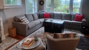 Ett vardagsrum med en grå hörnsoffa, ett litet runt bord med böcker, en korg av rotting och en dekorativ krans på väggen.