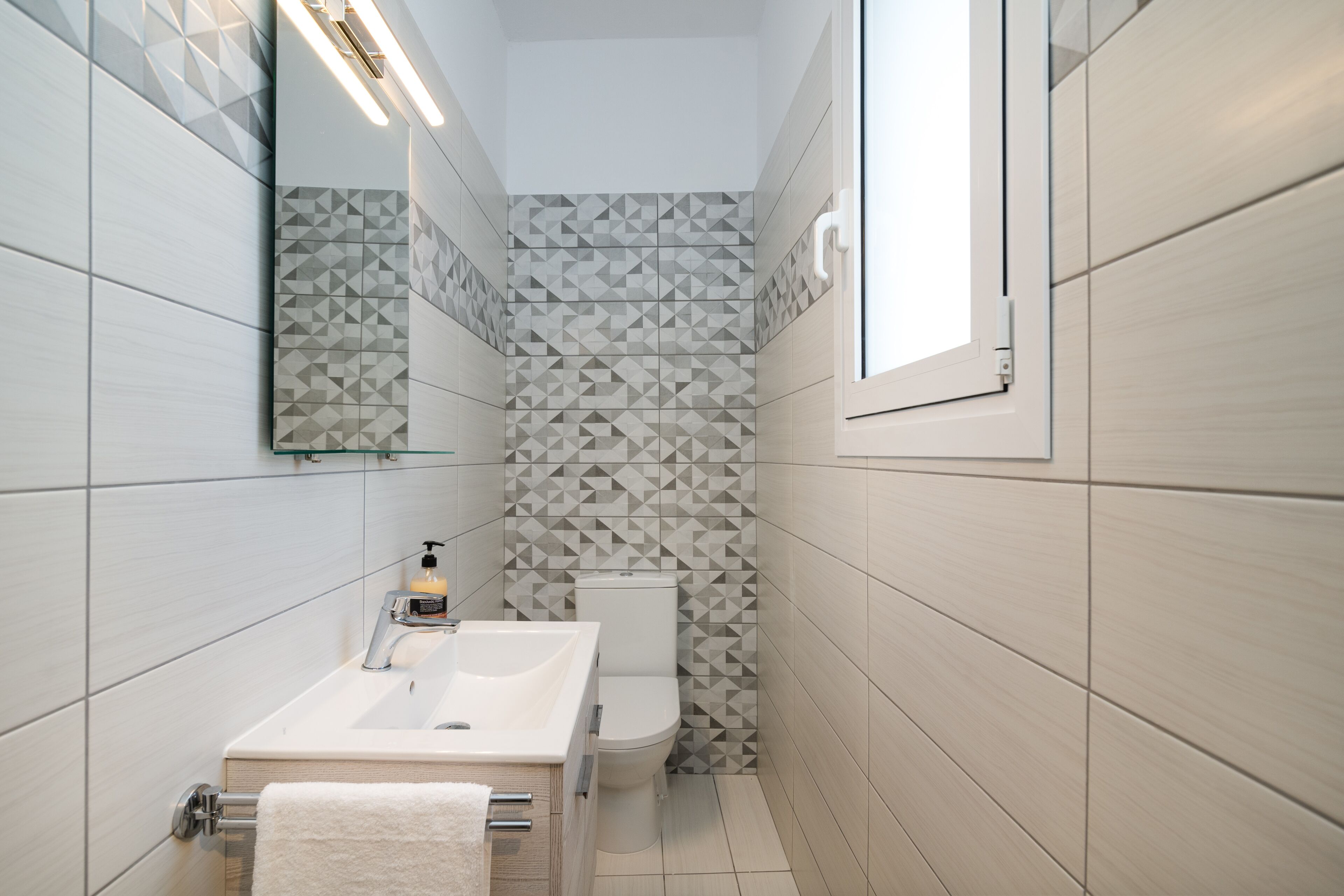 A bathroom with a white sink, mirror, and toilet. The walls have a geometric tile pattern.