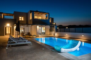 Un área de piscina moderna con sillones de descanso y un aro flotante, rodeada por un edificio de dos pisos con iluminación exterior.