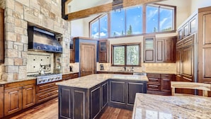A spacious kitchen with a large island, stone walls, wooden cabinets, and a high ceiling with exposed beams.
