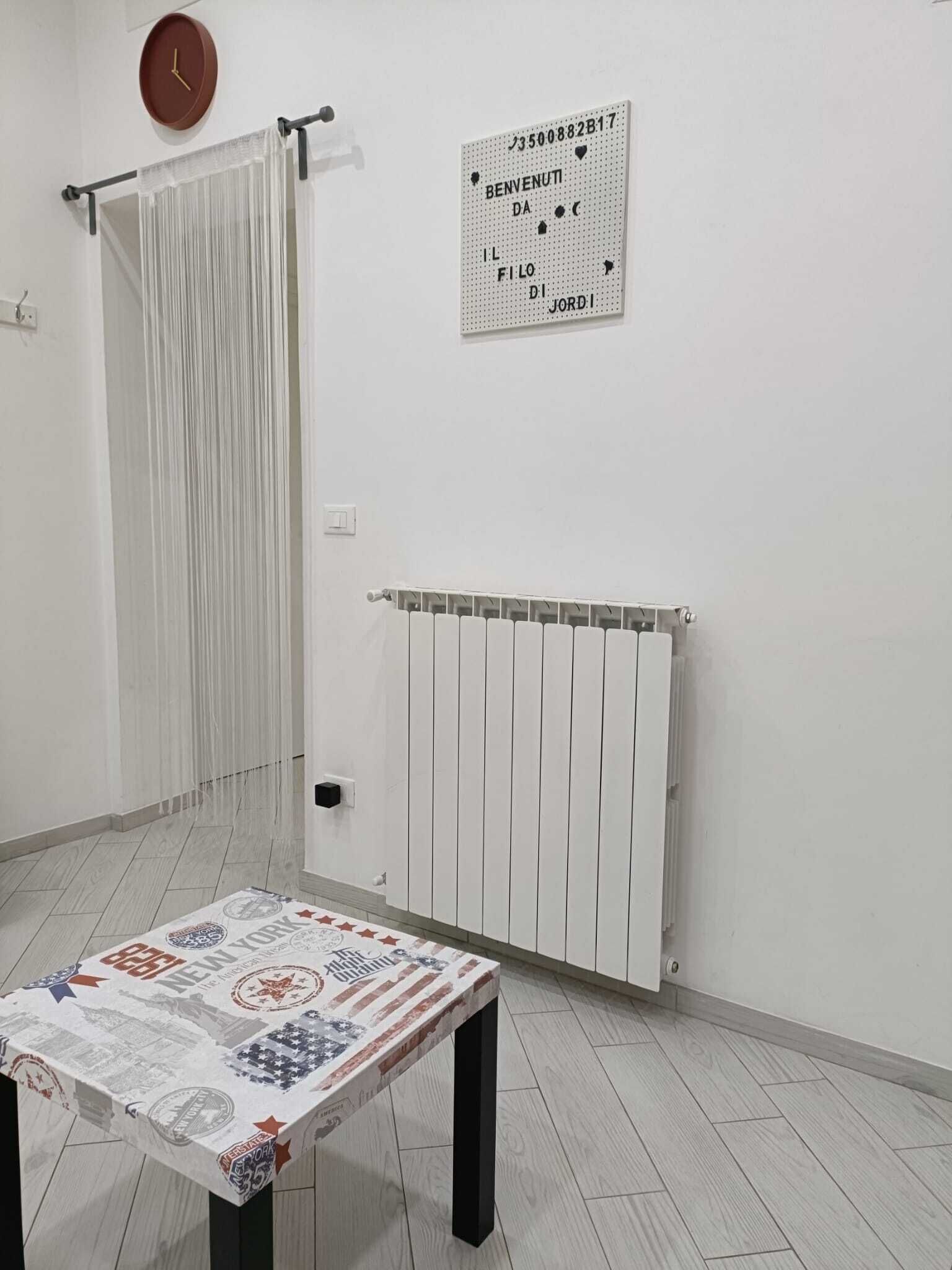 Ein Zimmer mit einem weißen Heizkörper, einem Tisch mit gemustertem Tischläufer, einem Vorhang und einem an der Wand befestigten Schild mit Text.
