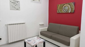 Une pièce avec un radiateur blanc, un canapé beige, une petite table recouverte d’une nappe à motifs, un mur d’accent rouge et un tableau abstrait encadré.