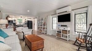 A spacious living room with a sofa, a wooden coffee table, and a rocking chair. There is a television mounted on the wall and a kitchen area in the background. - New Remodel! Updated Cottage near Beach and Park (Manistee)