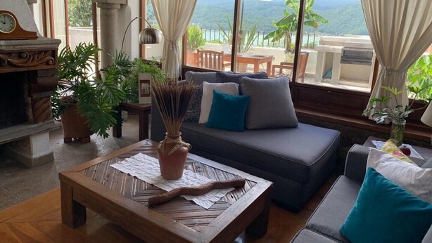 Uma sala de estar aconchegante com um sofá, uma mesa de centro e uma mesinha de cabeceira de madeira. Há uma janela com cortinas, um relógio e vista para o exterior.