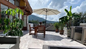 Um pátio externo com mesa, cadeiras, plantas em vasos e um grande guarda-sol.