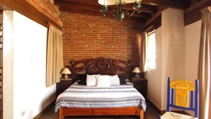 Quarto com parede de tijolos, teto de madeira, cama com cabeceira, duas mesas de cabeceira com luminárias, uma cadeira e uma janela com cortinas.