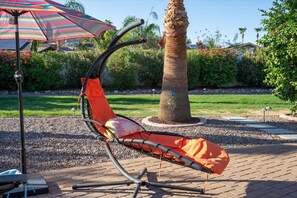 A hammock chair with an orange cushion, a striped umbrella, and a palm tree in a landscaped garden. - Home Theater | Private Pool | Hot Tub | Mini Golf (Scottsdale)