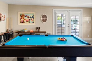 A pool table with a single white ball, surrounded by colored balls, in a room with a door, a framed painting of cows, and a wall-mounted sign.